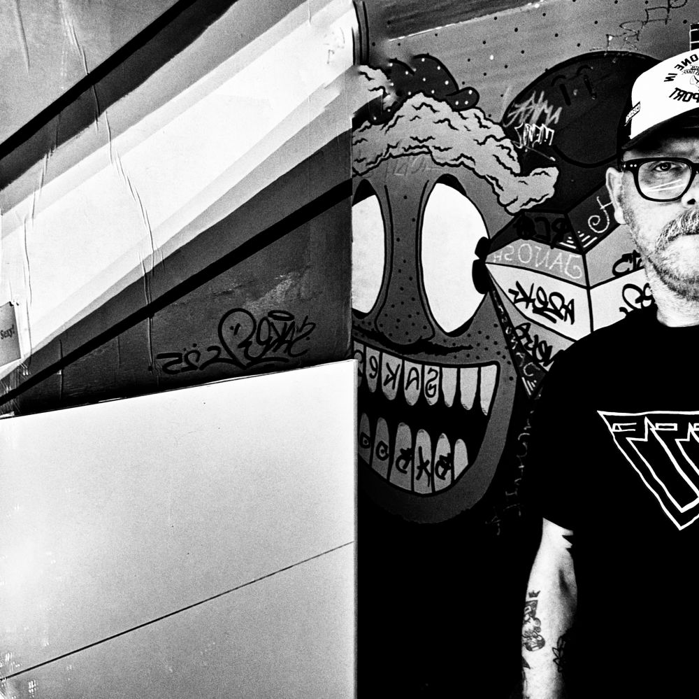 Ein Schwarz-Weiß-Foto von mir, auf dem ich neben einer Wand mit Cartoon-Graffiti stehe. Ich trage ein Basecap, eine Brille und ein schwarzes T-Shirt mit dem Logo der großartigen Punk-Musikgruppe »Pisse« auf der Vorderseite. Das Graffiti zeigt ein großes grinsendes Gesicht und andere abstrakte Bilder.